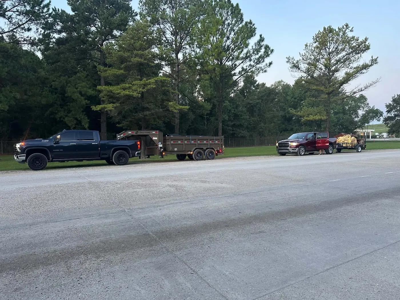 Two pickup trucks with junk removal trailers beside a wooded property