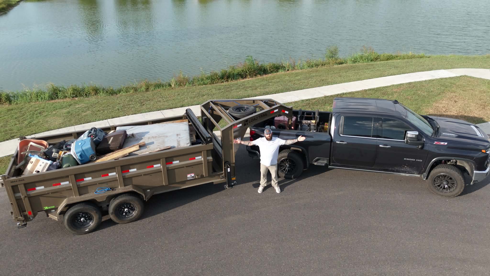 Loaded junk removal trailer hitched to a pickup truck beside a pond